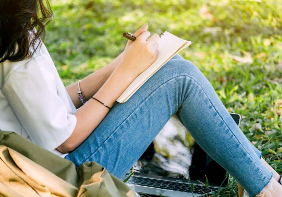 Woman with pen writing on a notebook sitting on grass in park