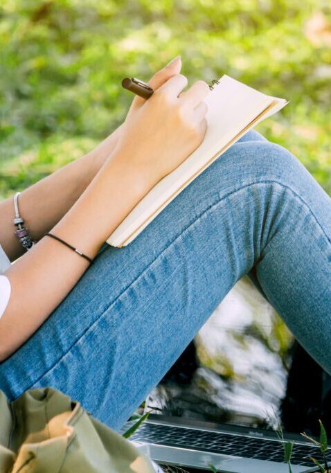 Person on grass writing in notebook, laptop nearby